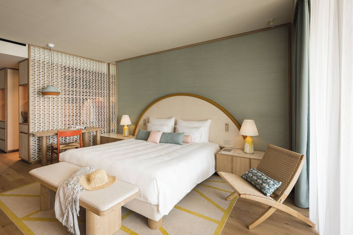 A hotel room with a large bed, a wooden bench, a chair, a small table, and a decorative wall panel.
