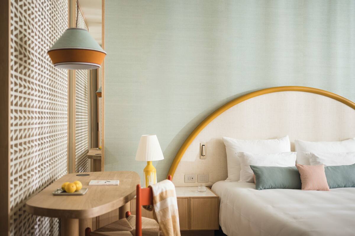 A hotel room with a wooden table, a lamp, a bed with pillows, and a decorative wall.