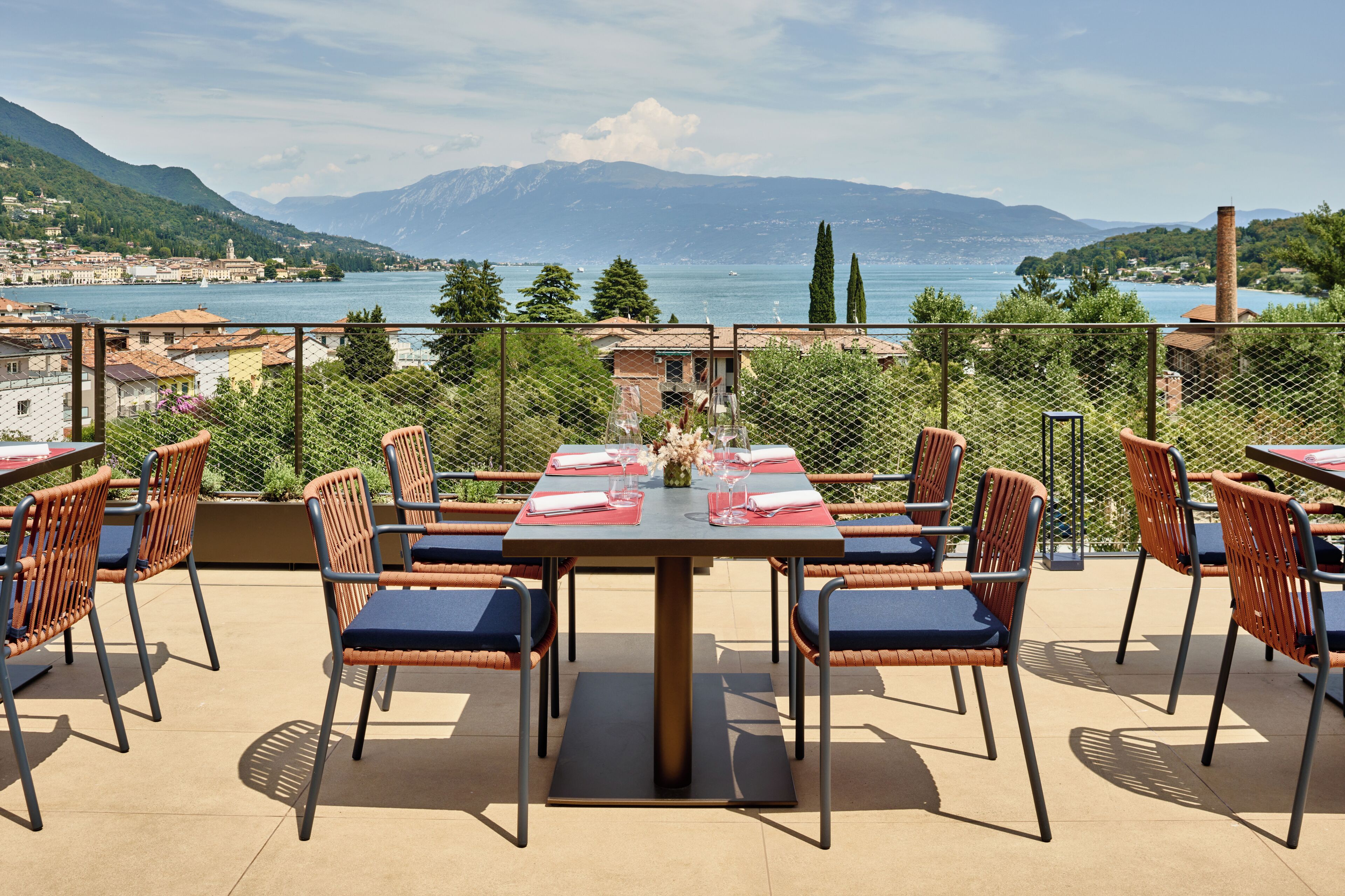 Un espacio de comedor al aire libre con una mesa lista para dos personas, rodeado de sillas de mimbre con cojines azules, con vistas a un lago y montañas panorámicos.