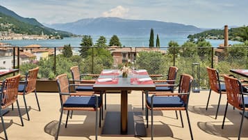 Un espacio de comedor al aire libre con una mesa lista para dos personas, rodeado de sillas de mimbre con cojines azules, con vistas a un lago y montañas panorámicos.