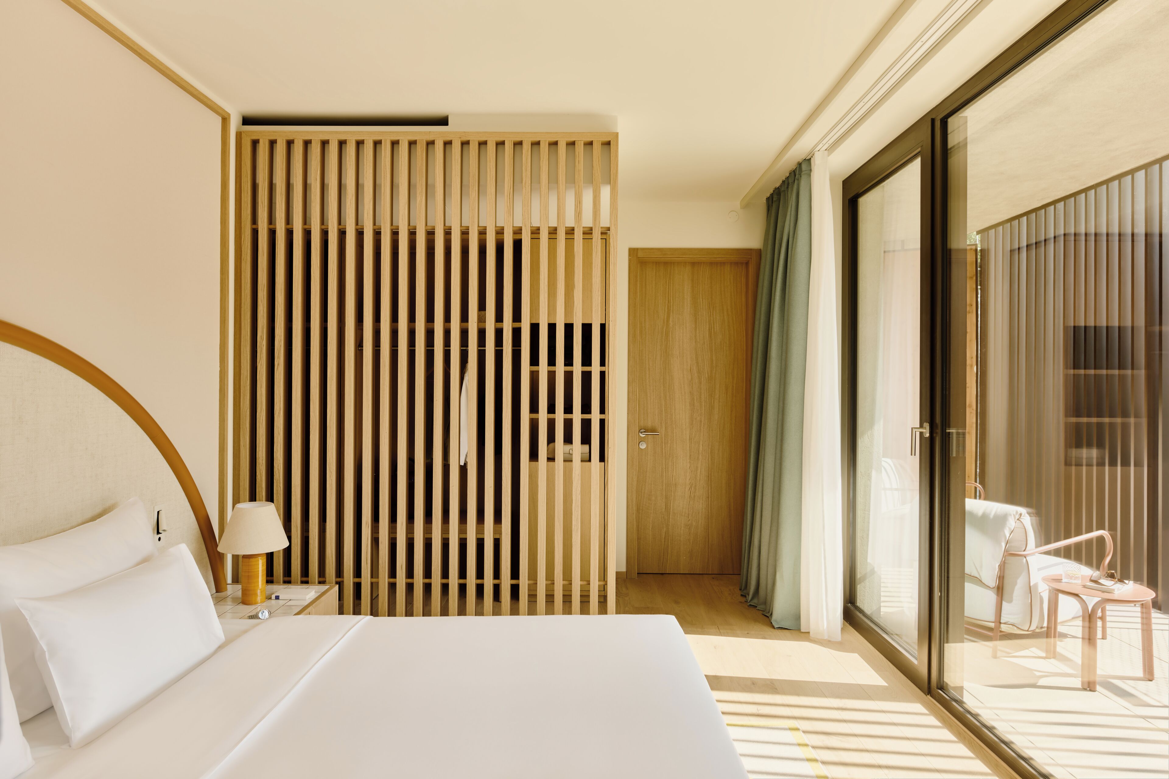 Habitación de hotel con una cama grande, una mesita de noche, una lámpara, una puerta corrediza de vidrio que da a un balcón y un panel de madera con una puerta.