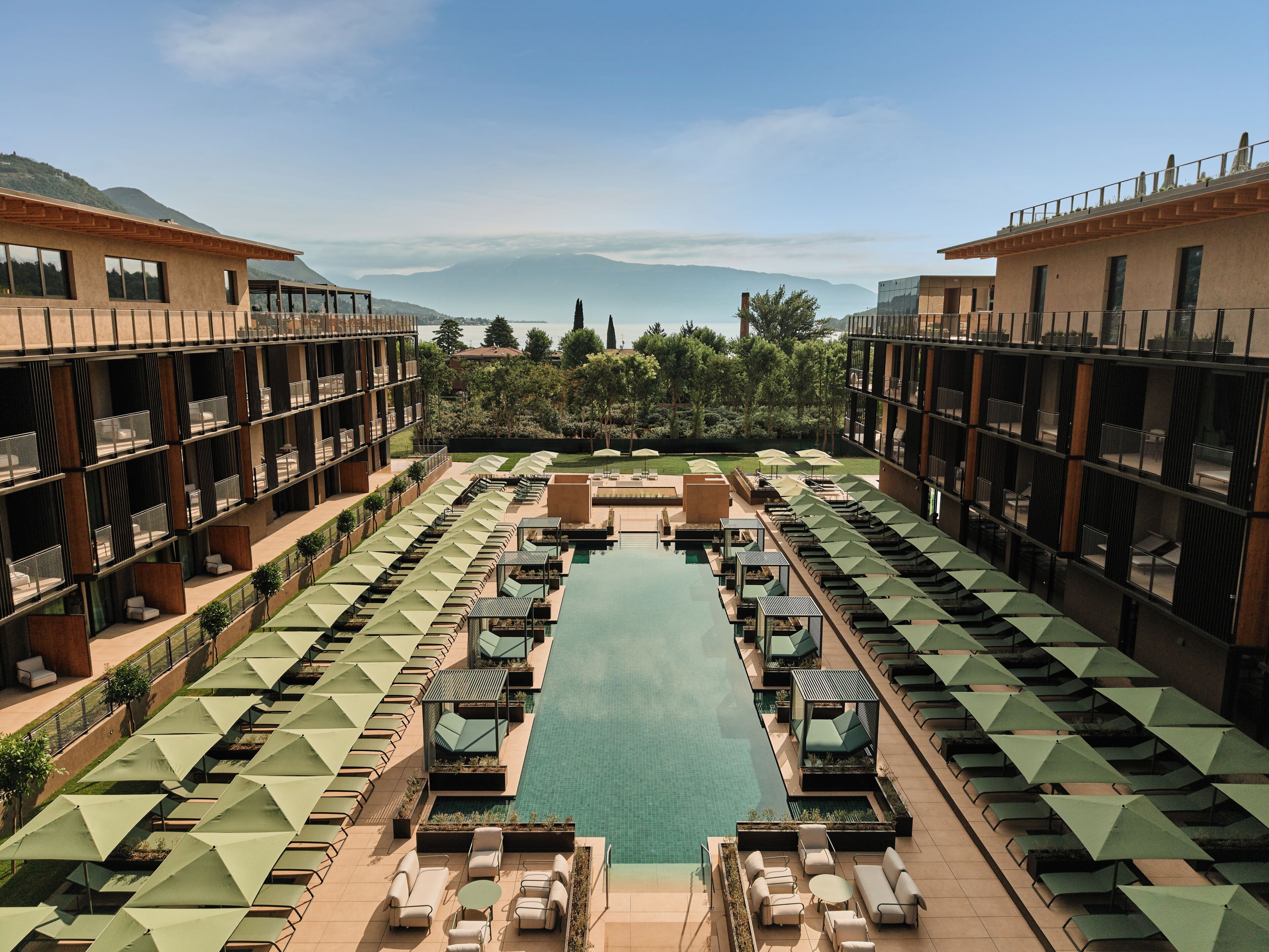Un hotel con piscina centrale, diverse zone relax con ombrelloni e vista sulle montagne.