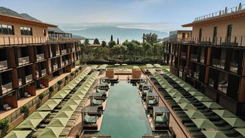 Un hotel con piscina centrale, diverse zone relax con ombrelloni e vista sulle montagne.