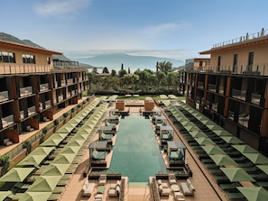 Un hotel con piscina central, varias zonas de descanso con sombrillas y vistas a las montañas.