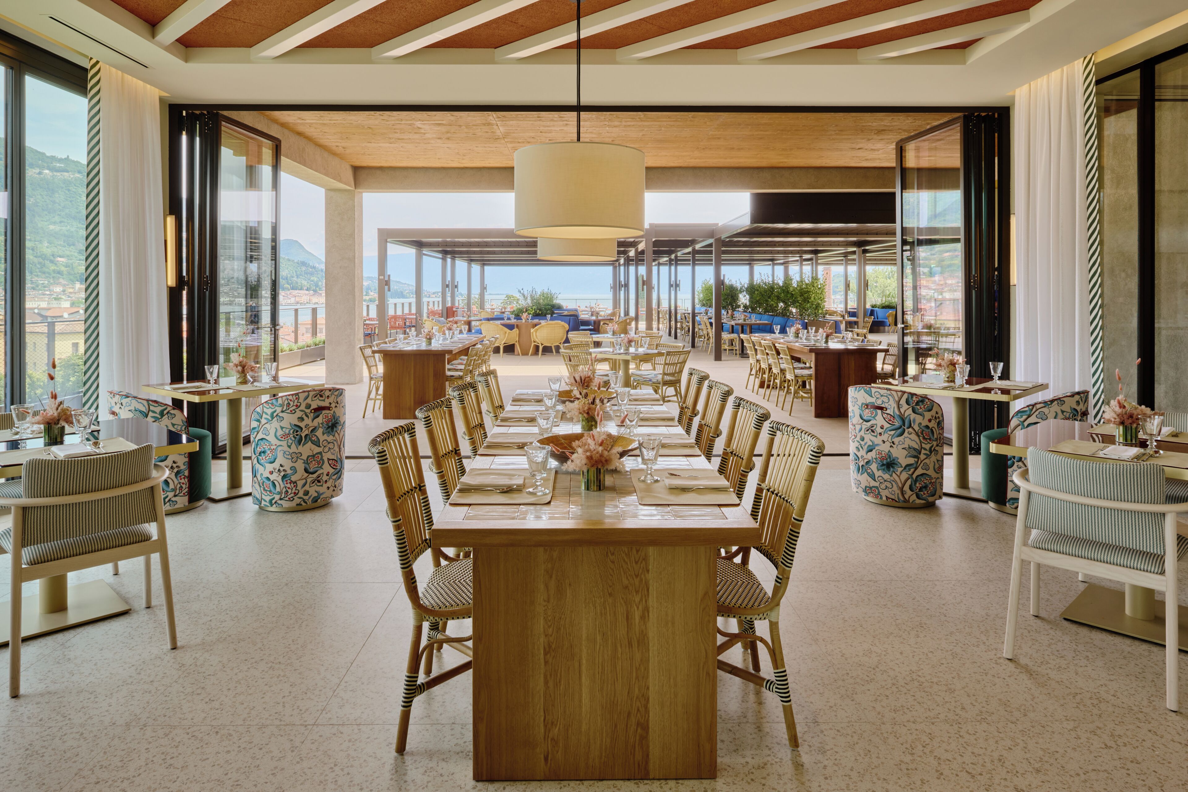 Un vaste espace dédié à la salle à manger, avec une longue table en bois et des chaises en osier, de grandes fenêtres offrant une vue sur la ville, et un plafond haut avec des poutres en bois.