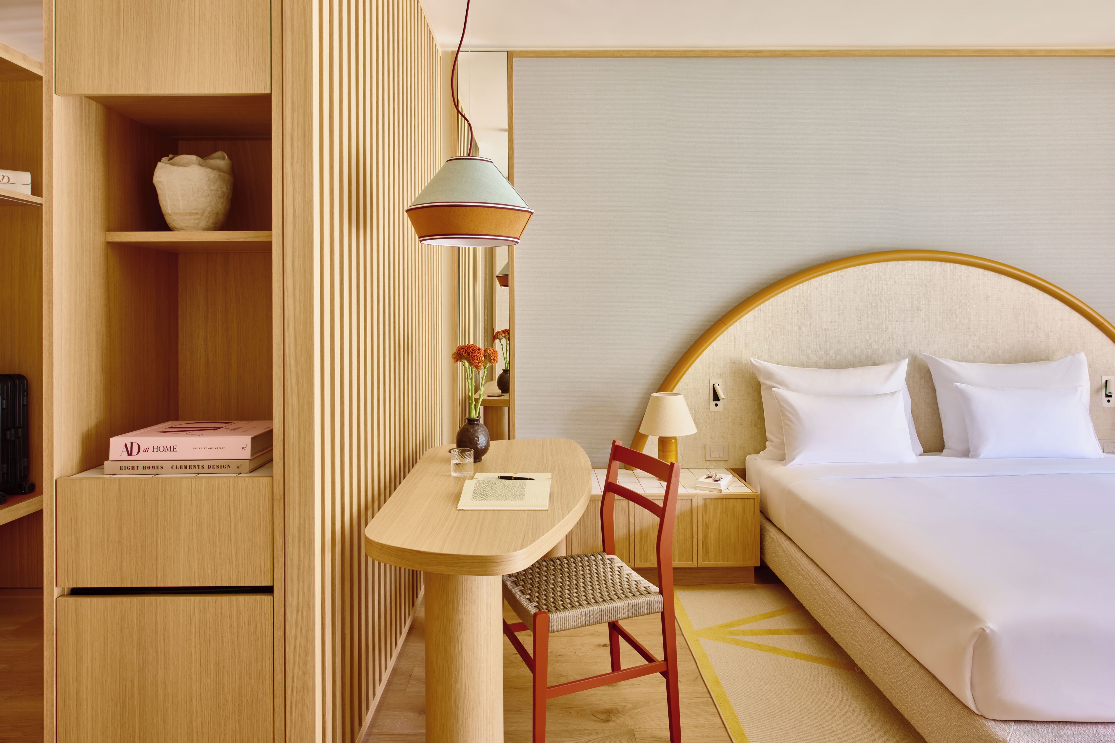 A hotel room with a bed, a desk with a chair, a lamp, a vase with flowers, and a bookshelf.