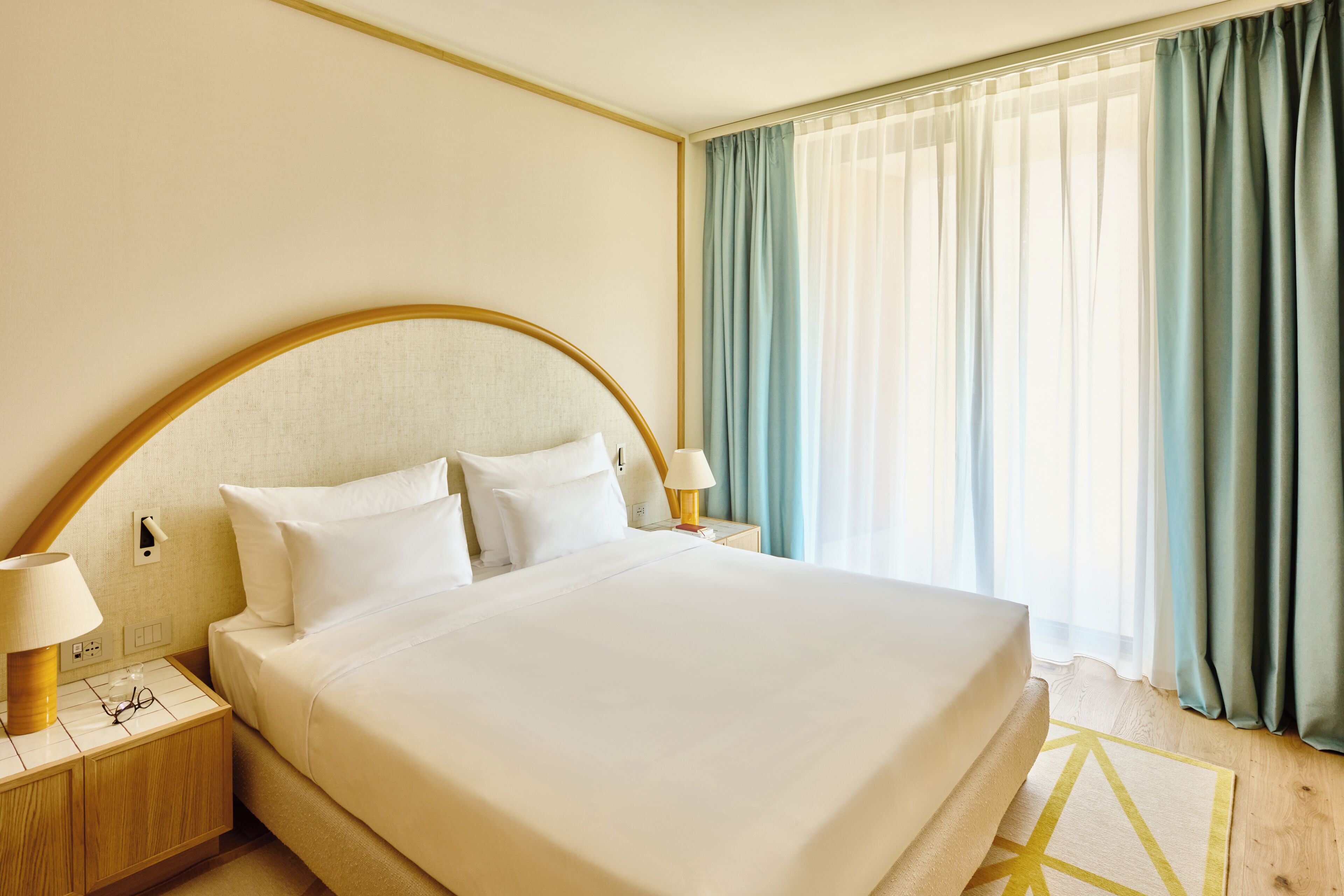 Una habitación de hotel con una cama grande, dos mesitas de noche, una lámpara y un ventanal con cortinas.