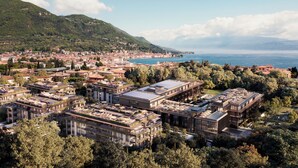 Vue aérienne d’une ville côtière, avec un mélange de bâtiments traditionnels et modernes, un port de plaisance et une chaîne de montagnes en arrière-plan.