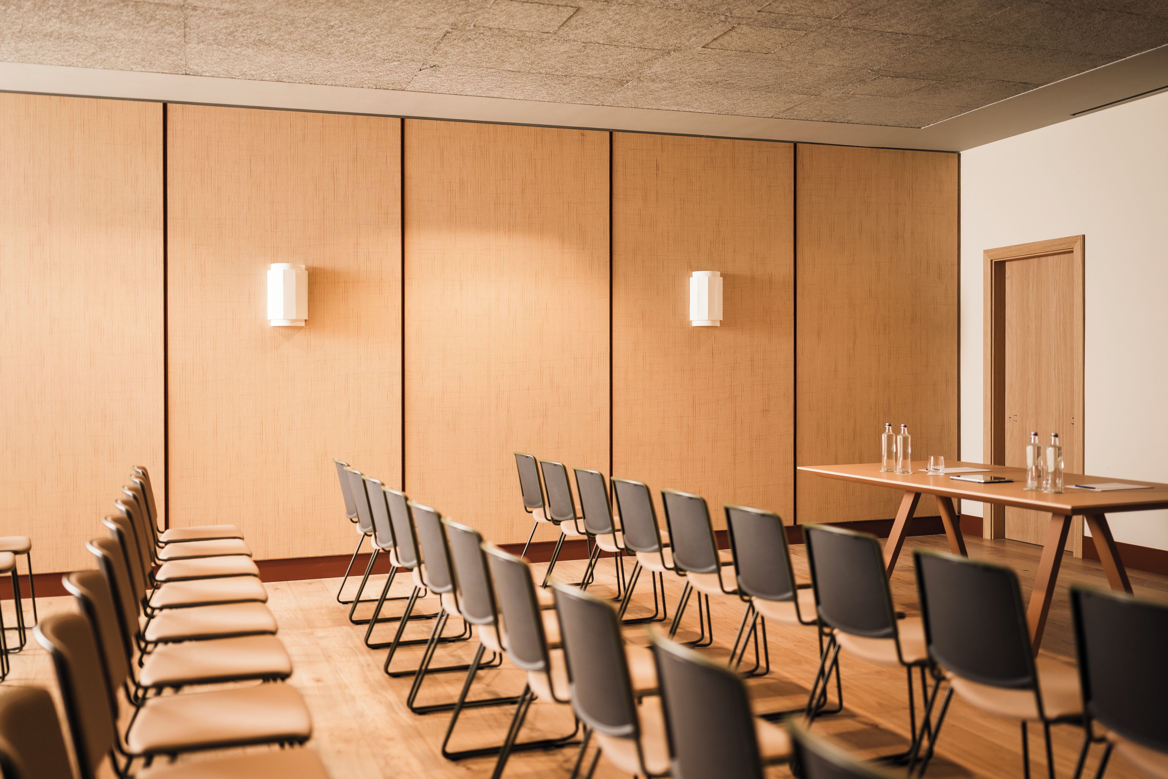 A conference room with a long table and rows of chairs, a door, and two wall-mounted light fixtures.