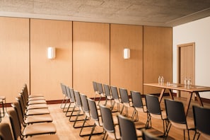 Un salón de conferencias con una larga mesa y filas de sillas, una puerta y dos luminarias fijadas a la pared.
