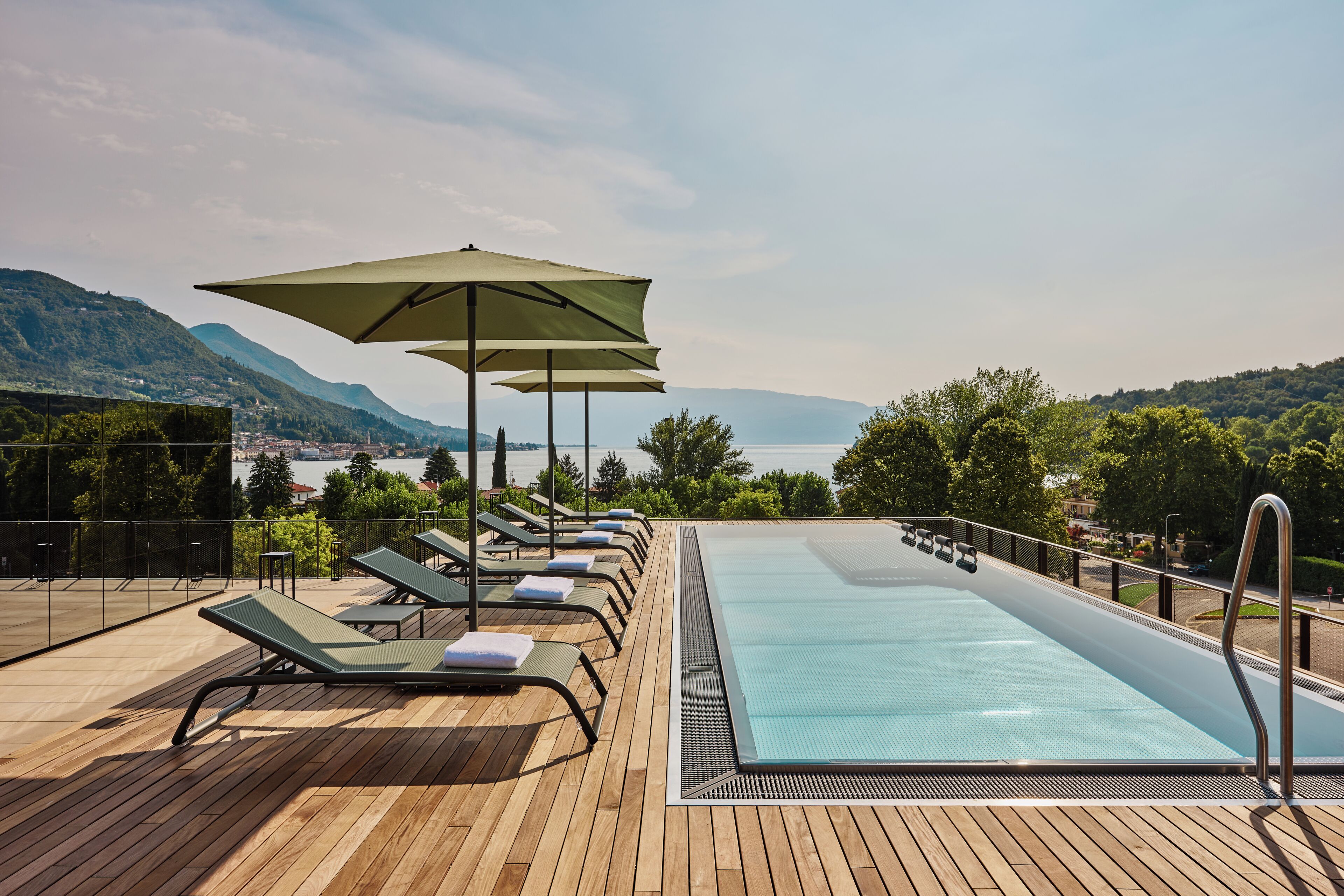 Zona junto a la piscina con sillas de descanso y sombrillas, con vistas a un cuerpo de agua y montañas.