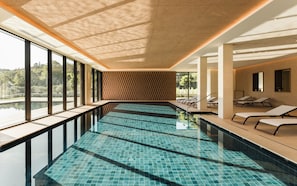 A modern indoor swimming pool with clear glass walls, a tiled floor, and lounge chairs along the sides. - Falkensteiner Resort Lake Garda (Salò)