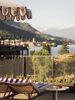 A terrace with lounge chairs, a table with a drink, and a view of a lake and mountains. - Falkensteiner Resort Lake Garda (Salò)