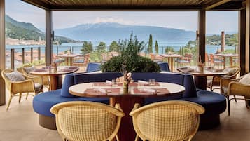 Een terras met ronde tafels en rieten stoelen, met uitzicht op een meer en bergen.