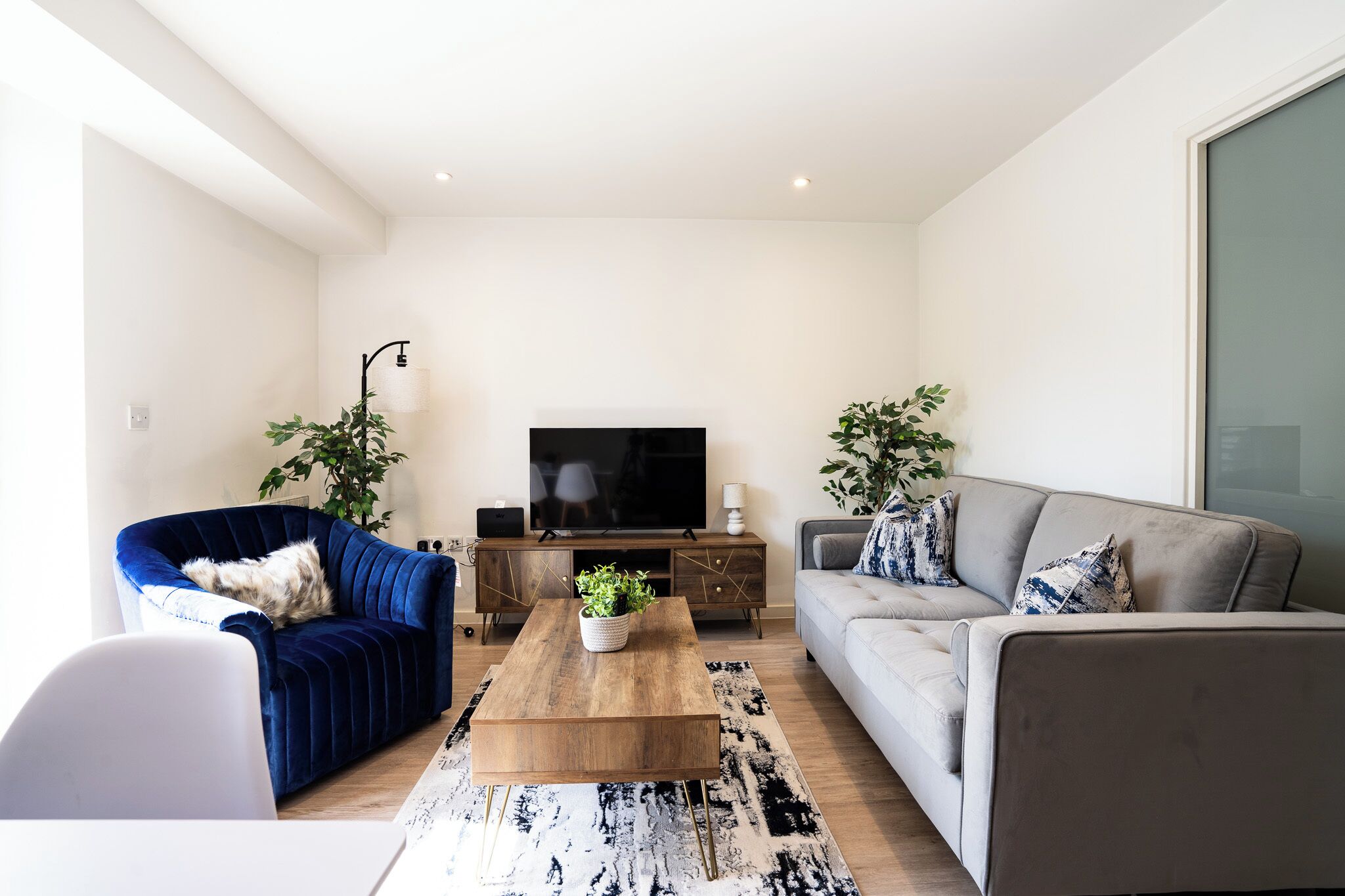 Un salon moderne avec un fauteuil bleu, un canapé gris, une table basse en bois et un téléviseur posé sur un meuble en bois.