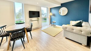 Un salon moderne comprenant un canapé, une table à manger avec des chaises, un téléviseur fixé au mur et une grande fenêtre offrant une vue sur l’extérieur.