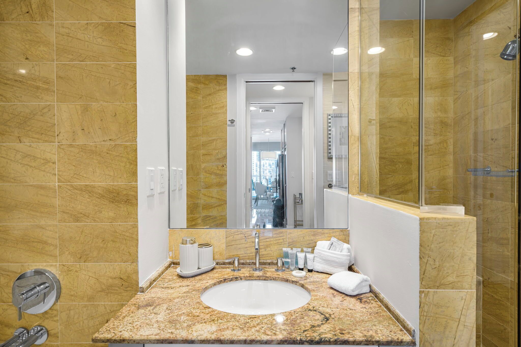 Une salle de bain moderne avec un lavabo surmonté d’un plan de travail en granit, un grand miroir et une cabine de douche en verre.