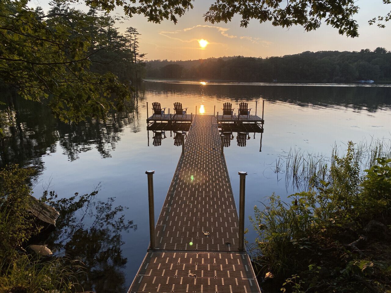 A wooden dock extending into a calm lake with chairs and tables at the end, surrounded by trees.
