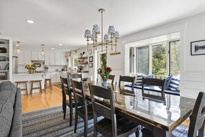 Een moderne keuken met witte kastjes, een eethoek met een grote houten tafel en uitzicht op de omgeving door grote ramen.