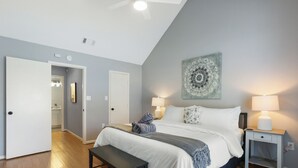 A bedroom with a bed, bedside tables, a bench, a ceiling fan, and a door leading to another room. - Adorable Bungalow Near Tybee Beach (Savannah)