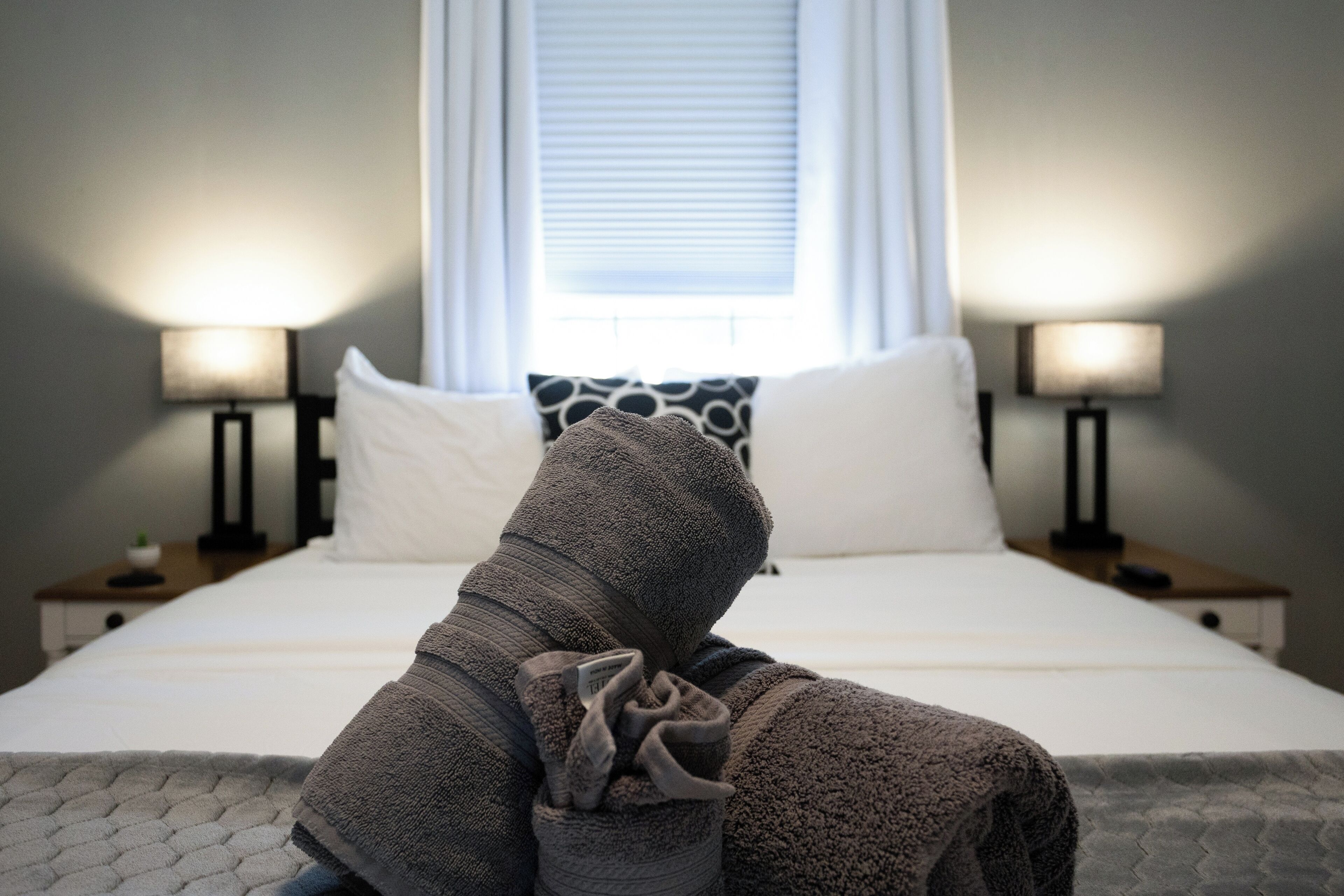 A neatly made bed with a white headboard, two bedside lamps, and a window with blinds.