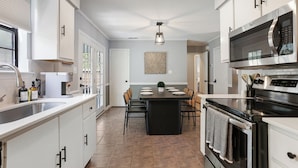 A modern kitchen with white cabinets, a central island, stainless steel appliances, and a dining area with chairs. - Adorable Bungalow Near Tybee Beach (Savannah)