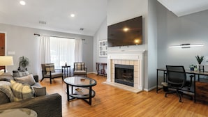 A spacious living room with a fireplace, a large flat-screen TV mounted above it, and a wooden floor. - Adorable Bungalow Near Tybee Beach (Savannah)
