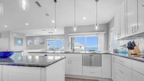 Cocina moderna con armarios blancos, una isla grande, electrodomésticos de acero inoxidable, iluminación colgante y vista al océano.