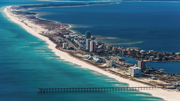 Vista aérea de una zona turística costera con una larga playa de arena, un muelle que se adentra en el océano y un conjunto de edificios a lo largo de la costa.