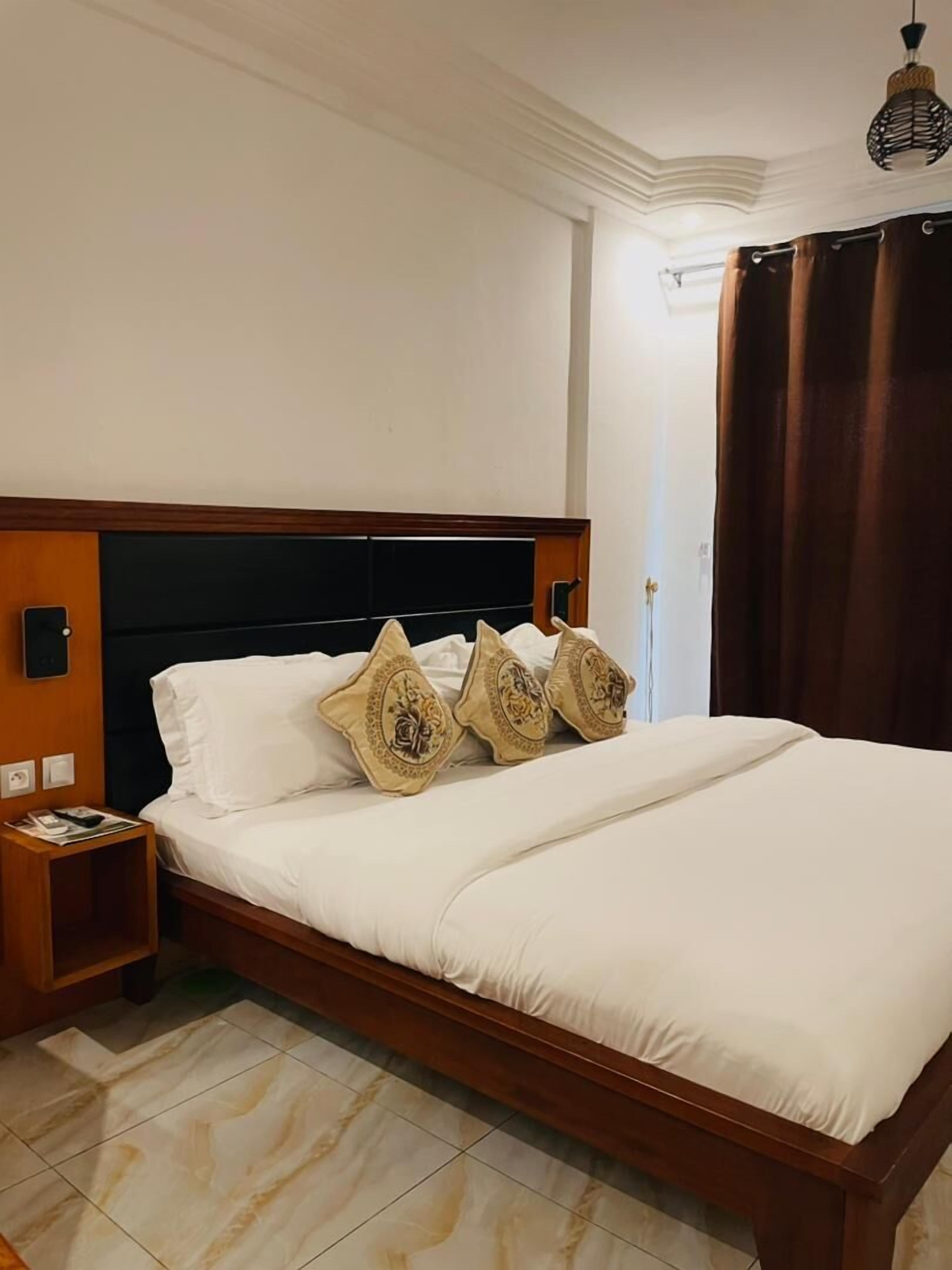 A neatly made bed with white linens and decorative pillows, a wooden headboard, a bedside table with a lamp, and a tiled floor.
