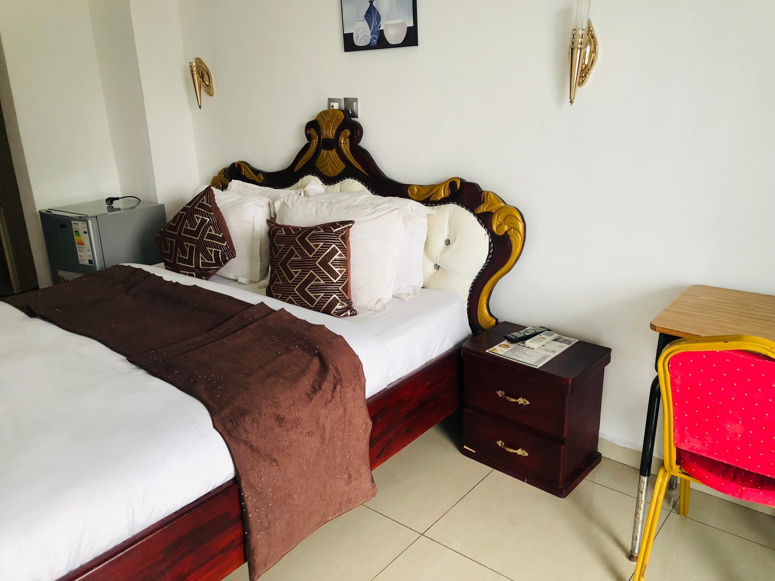 A hotel room with a large bed, a bedside table with drawers, a small desk with a chair, and a wall-mounted lamp.