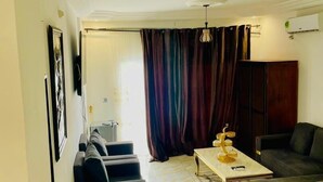 A living room with a sofa, a coffee table, and a TV. There is a curtain and a window. - Résidence hôtelière Samba (Douala)