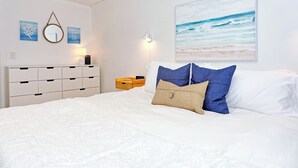 Une chambre à coucher bien rangée, avec un lit, une commode, un miroir fixé au mur et un tableau sur le thème de la plage.