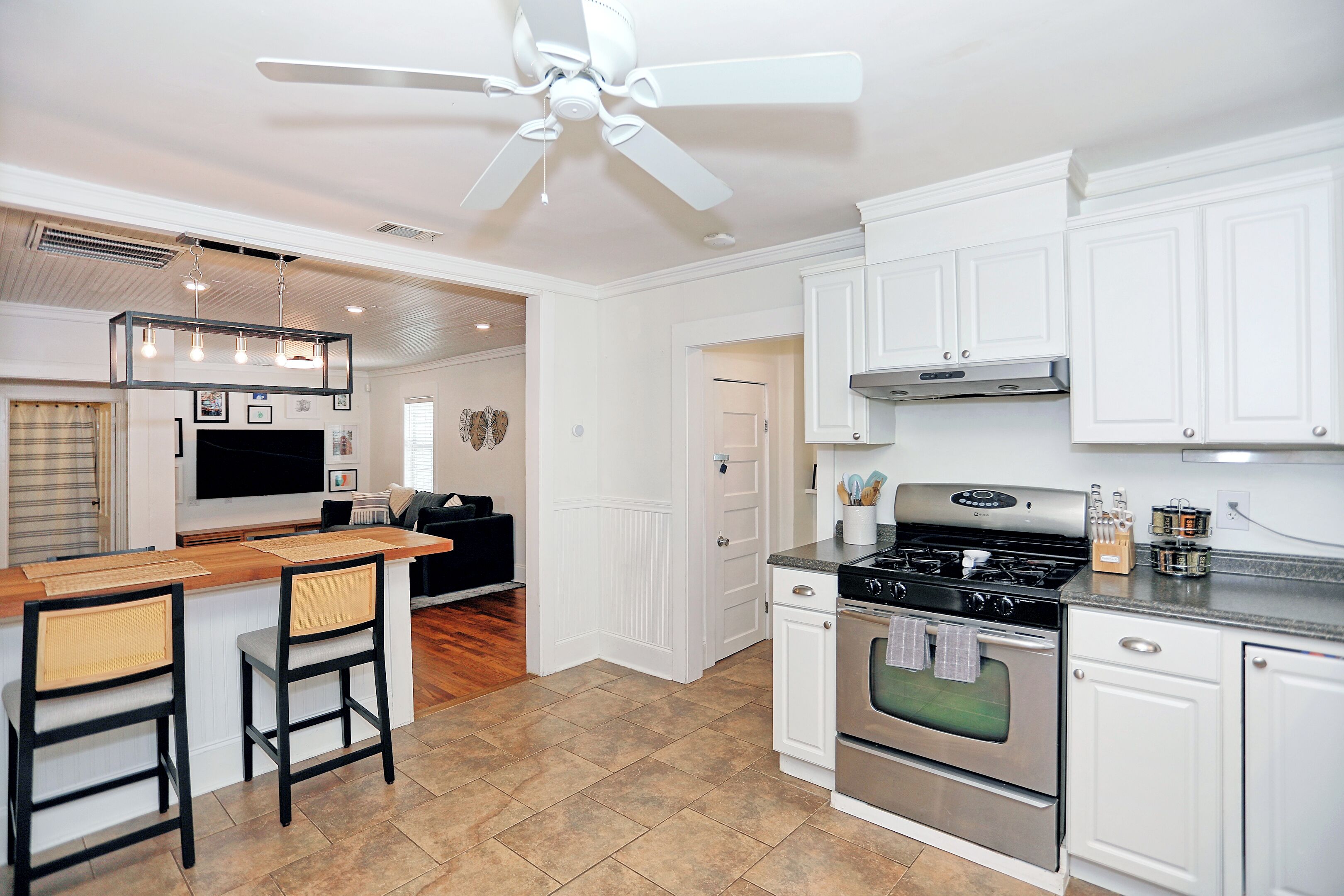 A modern kitchen with white cabinetry, a central island with bar stools, a stainless steel stove, and a ceiling fan.