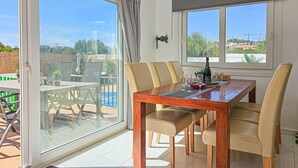Espaço de refeições com mesa de madeira, cadeiras bege e vista para uma piscina e vegetação lá fora.