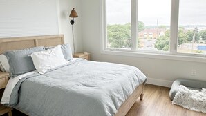 Une chambre avec un grand lit, une table de chevet, une lampe, une fenêtre donnant sur des immeubles et un petit tapis.
