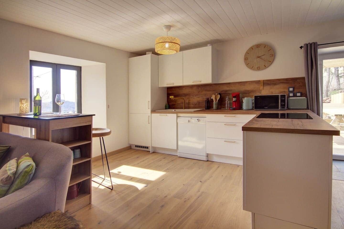 A modern kitchen with white cabinets, a wooden countertop, and a built-in dishwasher. There is a dining area with a table and chairs, and a large window providing natural light.