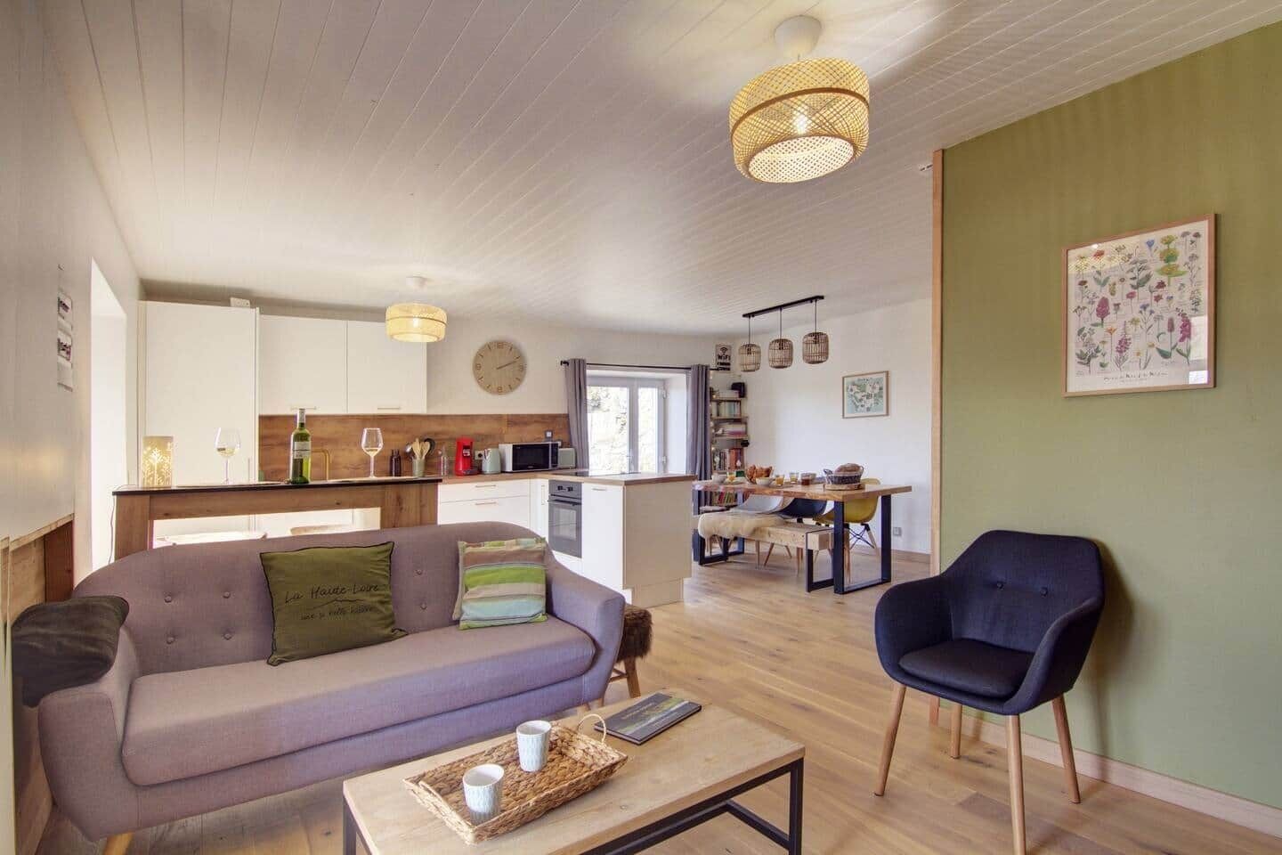 A cozy living room with a sofa, a coffee table, and a chair. A kitchen with wooden cabinets and a dining area with a table and chairs is visible in the background.