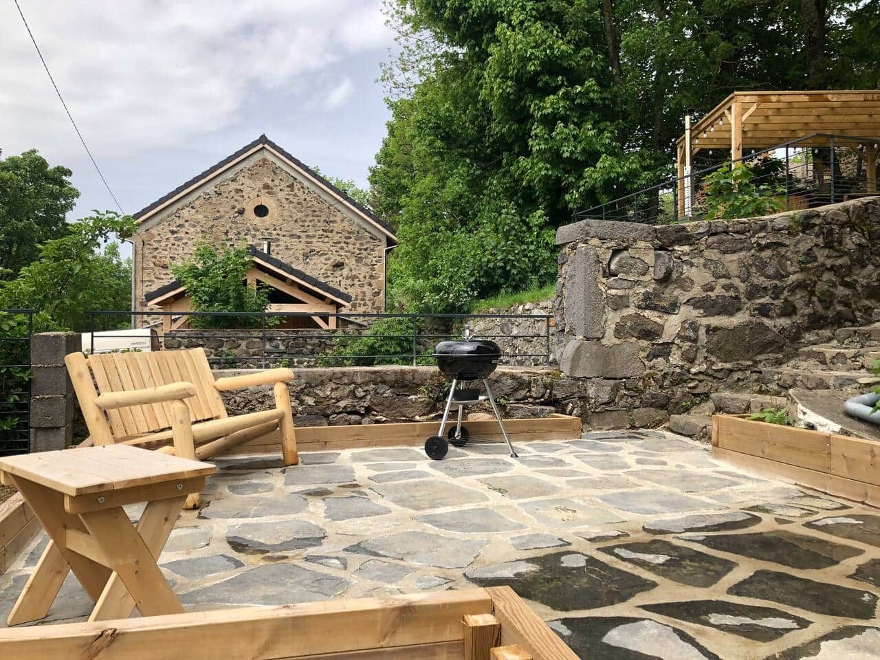 Een terras van natuursteen met een houten barbecue, een houten bank en een tafel.