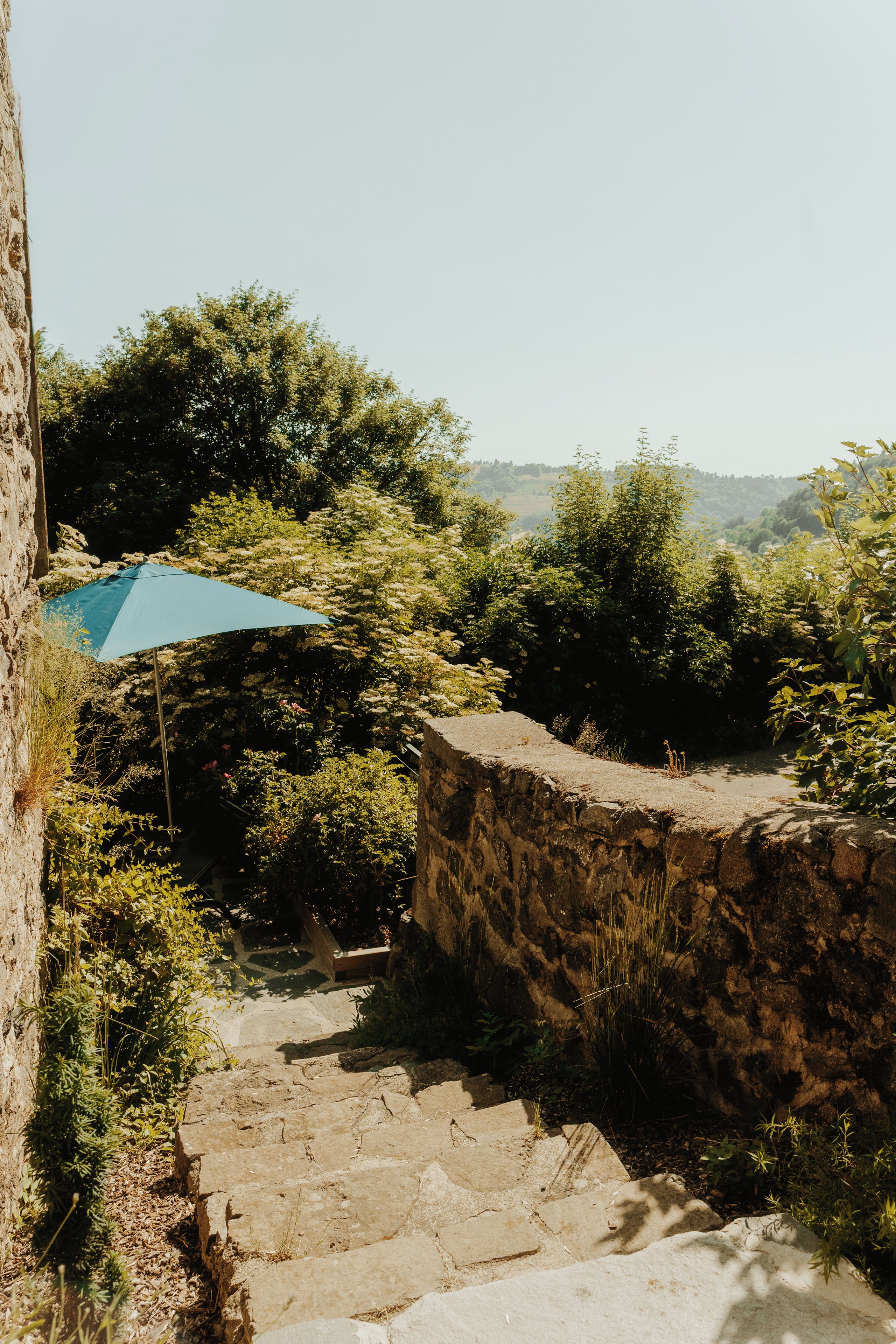 Un escalier en pierre mène à un parasol bleu, entouré d’une végétation luxuriante et d’un mur de pierre.
