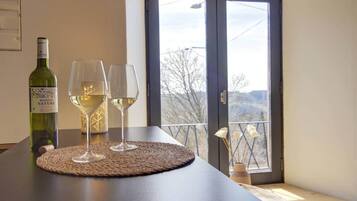 Ein Tisch mit einer Weinflasche und zwei Gläsern auf einer gewebten Tischunterlage, ein Fenster mit Blick auf Bäume und Hügel sowie eine Tür mit Griff.