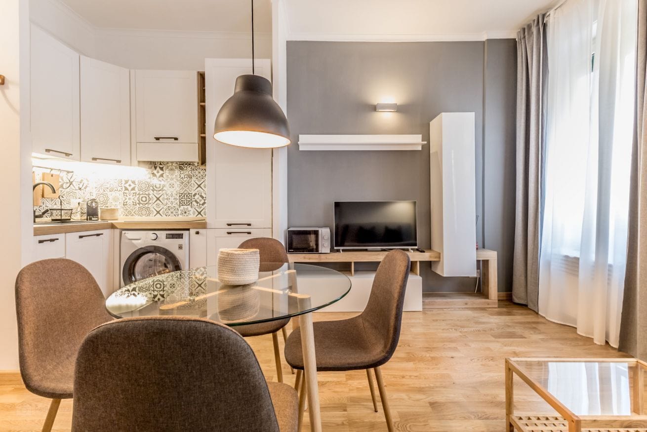 Eine moderne Küche mit Mikrowelle, Toaster und Waschmaschine. Ein Essbereich mit Glastisch und Stühlen ist vorhanden. Ein Fernseher ist an der Wand montiert, darüber hängt eine große Pendelleuchte.