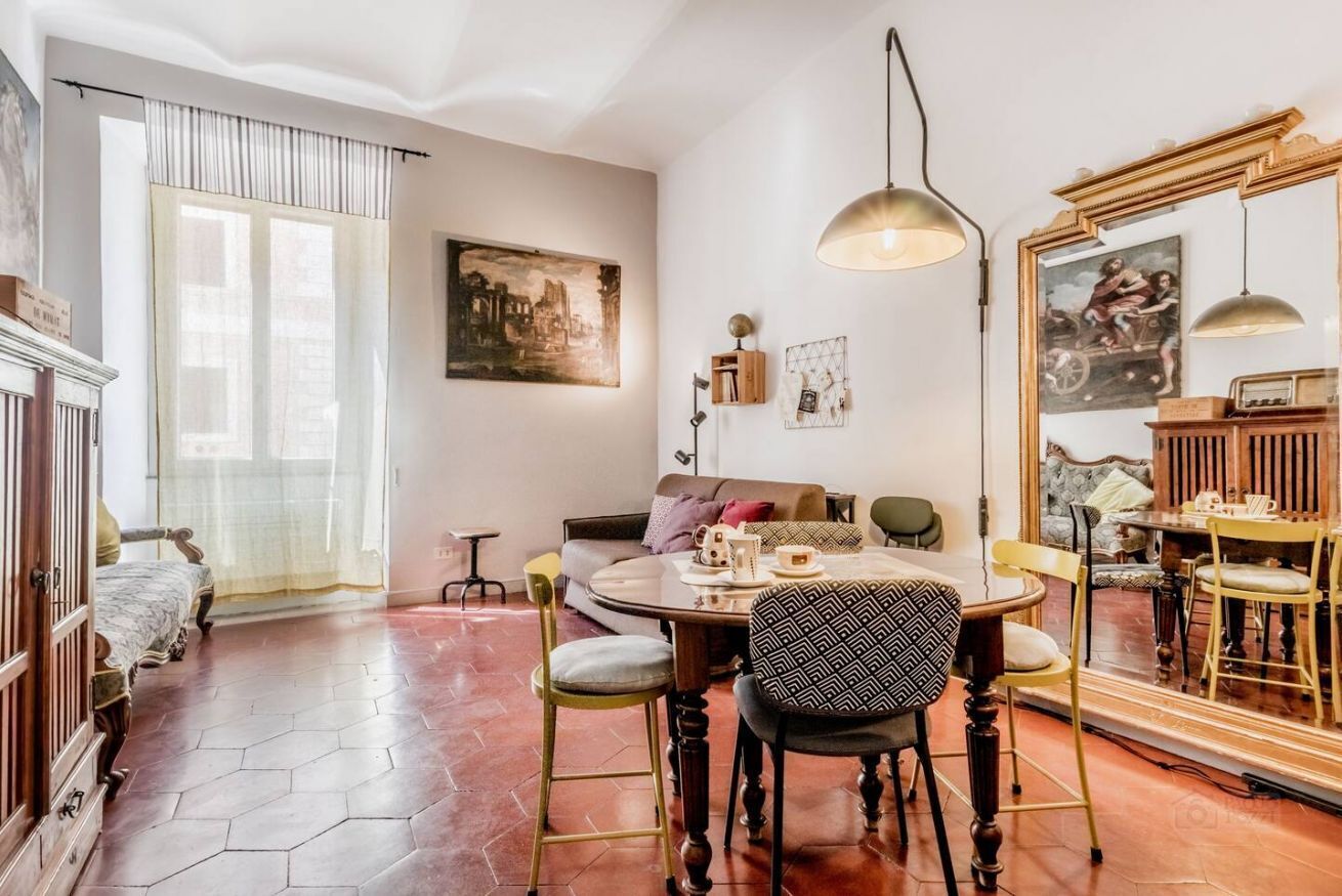 A cozy living room with a round wooden table, comfortable seating, and a large mirror reflecting the room.