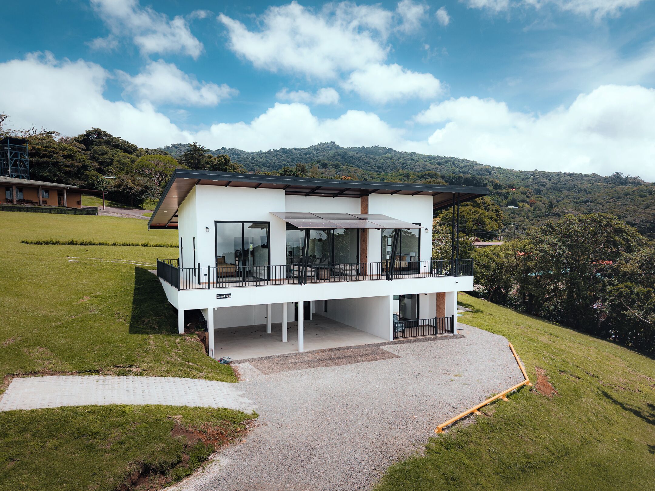 A modern two-story house with a balcony, large windows, and a view of the surrounding hills.