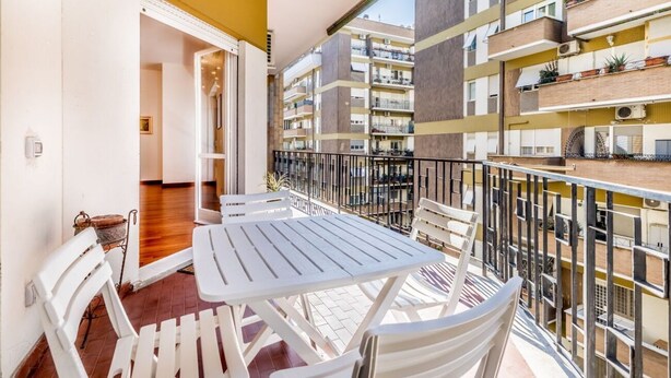 Un balcón con mesas y sillas blancas, que da a un edificio residencial de varios pisos con balcones.