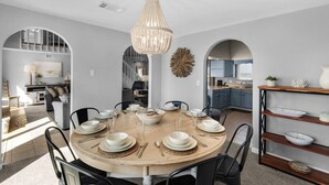 A dining area with a round table set for six, surrounded by chairs, with a chandelier above.