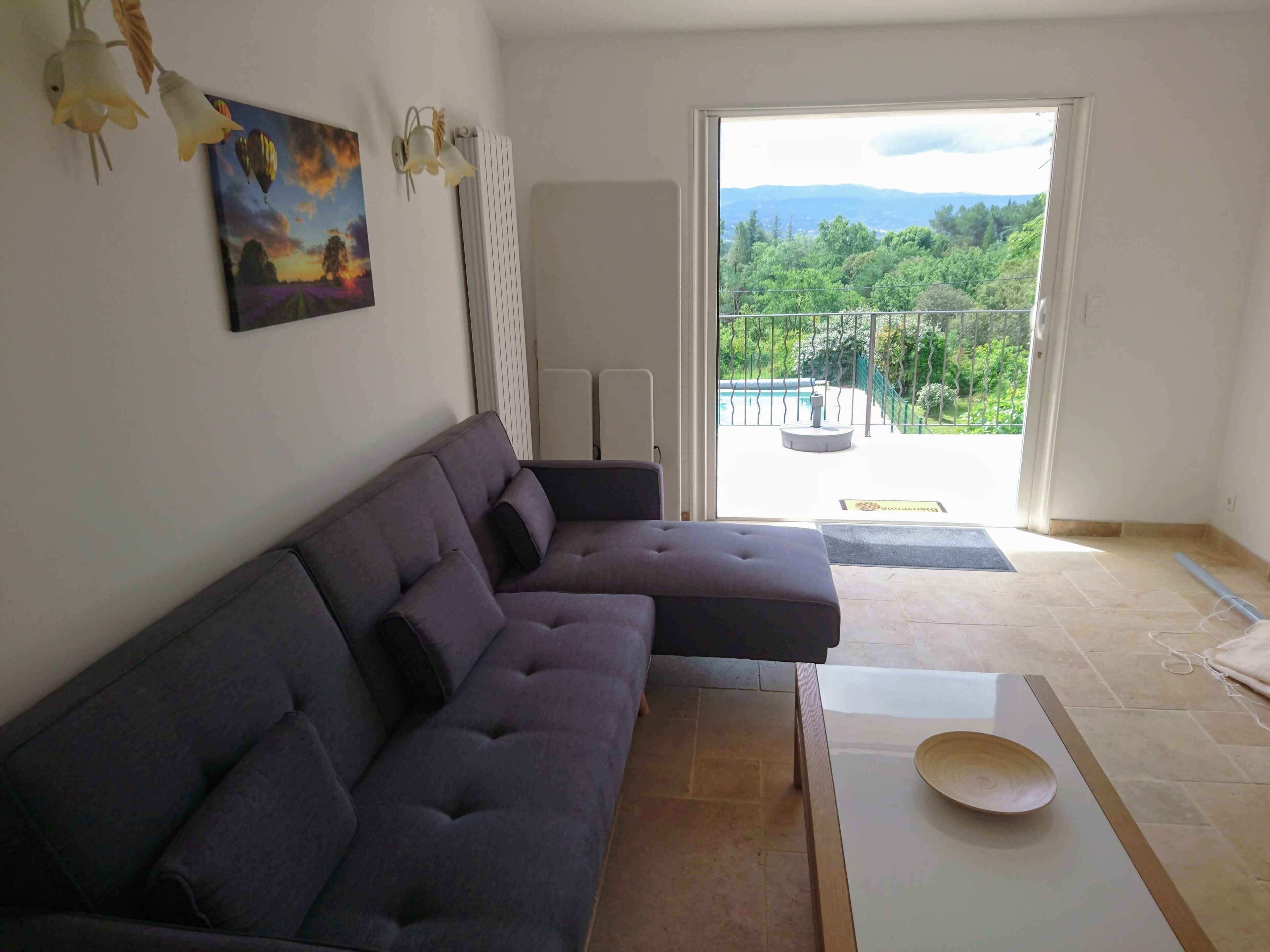 Un salon moderne avec un canapé d’angle, une table à manger agrémentée d’un saladier, un tableau et une vue sur un balcon verdoyant.