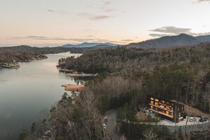 A modern house with a large deck overlooking a river and forested hills.