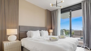 A modern hotel room with a large bed, two bedside tables with lamps, a view of the city through large windows, and a cityscape visible outside. - Calanova Escape | terrace - pool - golf (La Cala de Mijas)