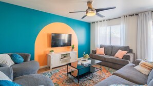 A living room with a teal accent wall, a ceiling fan, a flat-screen TV, a white entertainment unit, and a gray sofa with orange cushions. - Sleeps 20! Movie Theater, Arcade & King Suites near Stadium (Glendale)